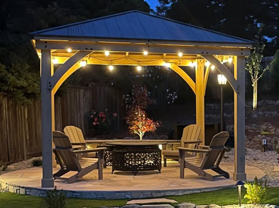 Illuminated wood pavilion with outdoor seating arranged around a fire pit